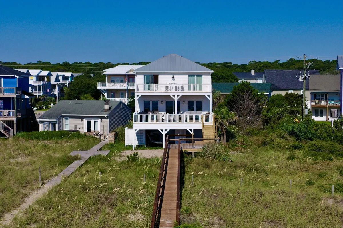 A wide exterior shot shows the beach house, its private wooden walkway, and the surrounding greenery leading toward the ocean. The house is inviting, with balconies and outdoor seating, reinforcing communal outdoor living and togetherness. The scene hints at sunrise or sunset, enhancing the sense of relaxation and shared memories.