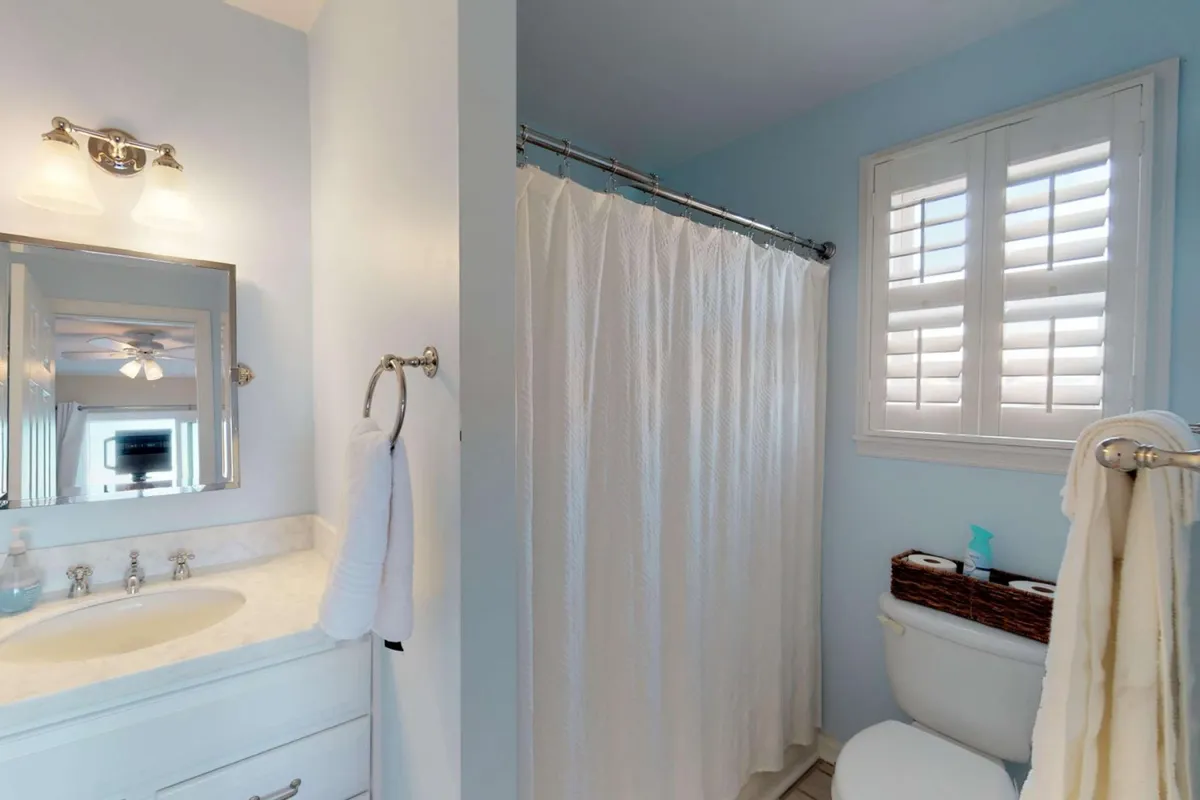 A tranquil bathroom scene featuring a vanity with a mirror, shower curtain, toilet, and a window with shutters. The space is clean and inviting, suggesting a stocked and well-maintained bathroom ideal for relaxing self-care routines.