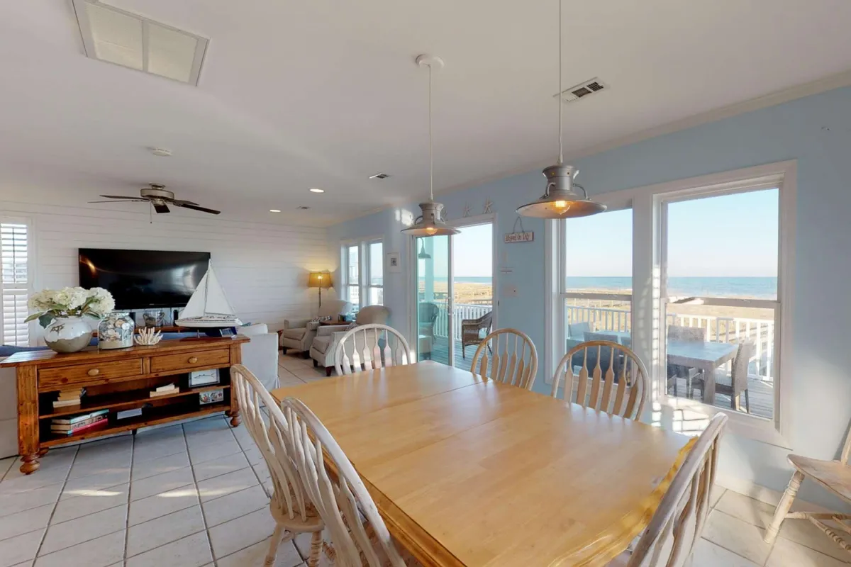 The main living and dining space featuring a large dining table positioned by expansive windows, comfortable seating, and a television. The room is bathed in natural light and offers clear views of the ocean, ideal for family meals, game nights, or relaxing while watching the sunset.
