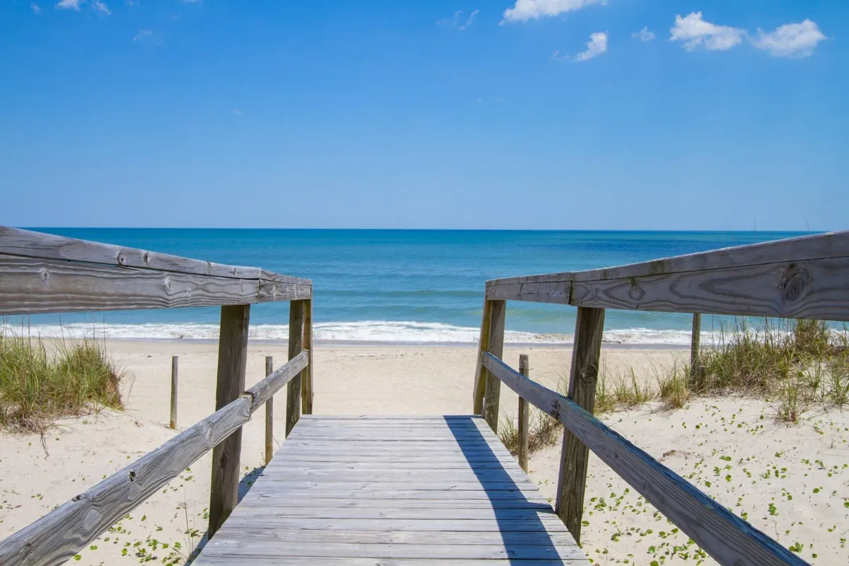 A wooden walkway extends directly from the property out to the sandy beach, with the ocean waves visible in the background. The scene captures the easy, direct access from the home to the shoreline, with footprints in the sand, gentle surf, and ample space for beach activities. The early morning or late afternoon light creates a relaxed, inviting mood.