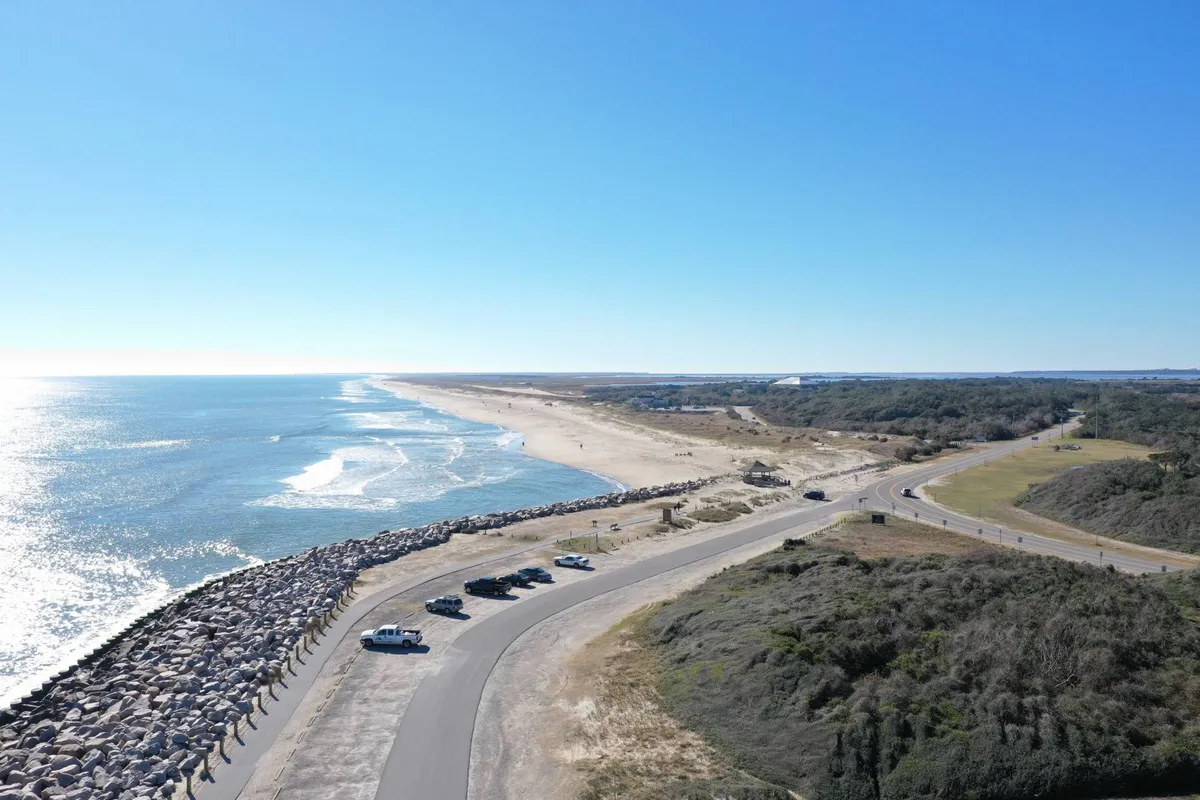 This image presents an inviting scene of the beach and ocean, with a jetty, vehicles parked nearby, and open sandy stretches perfect for walking and relaxing. The mood is relaxed, showing both human and natural elements harmoniously coexisting.