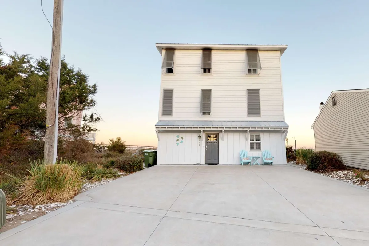 A three-story vacation home with blue chairs on the porch and a beautifully landscaped garden in front. The inviting exterior suggests plenty of space for group activities and captures the home's welcoming, family-friendly atmosphere.