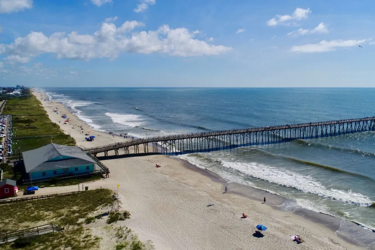A dynamic exterior scene featuring the Kure Beach Pier jutting into the ocean, with waves rolling onto the sandy shore and people enjoying various beach activities. The pier is bustling, and the beach is lively with families and groups sunbathing, swimming, and building sandcastles.