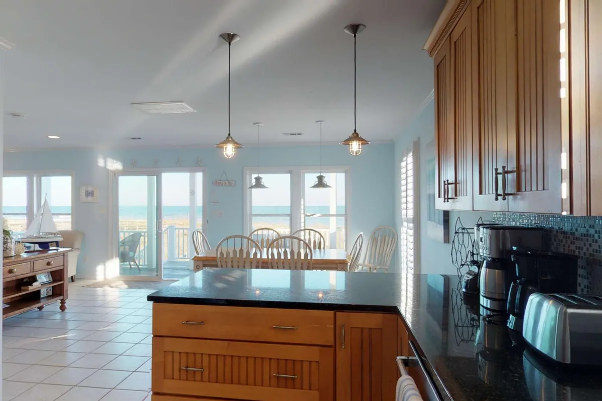 This image showcases a stylish, fully equipped kitchen with a granite island countertop, adjacent dining area, and large windows offering an ocean view. The setup is inviting for group cooking, family meals, and entertaining.