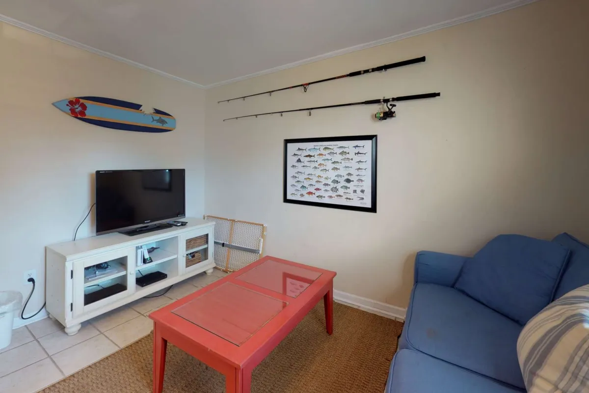 A cozy interior living space featuring a television, red coffee table, blue couch, and fishing rods on the wall. The playful fish illustration and relaxed seating make it ideal for gatherings, games, or movie nights.