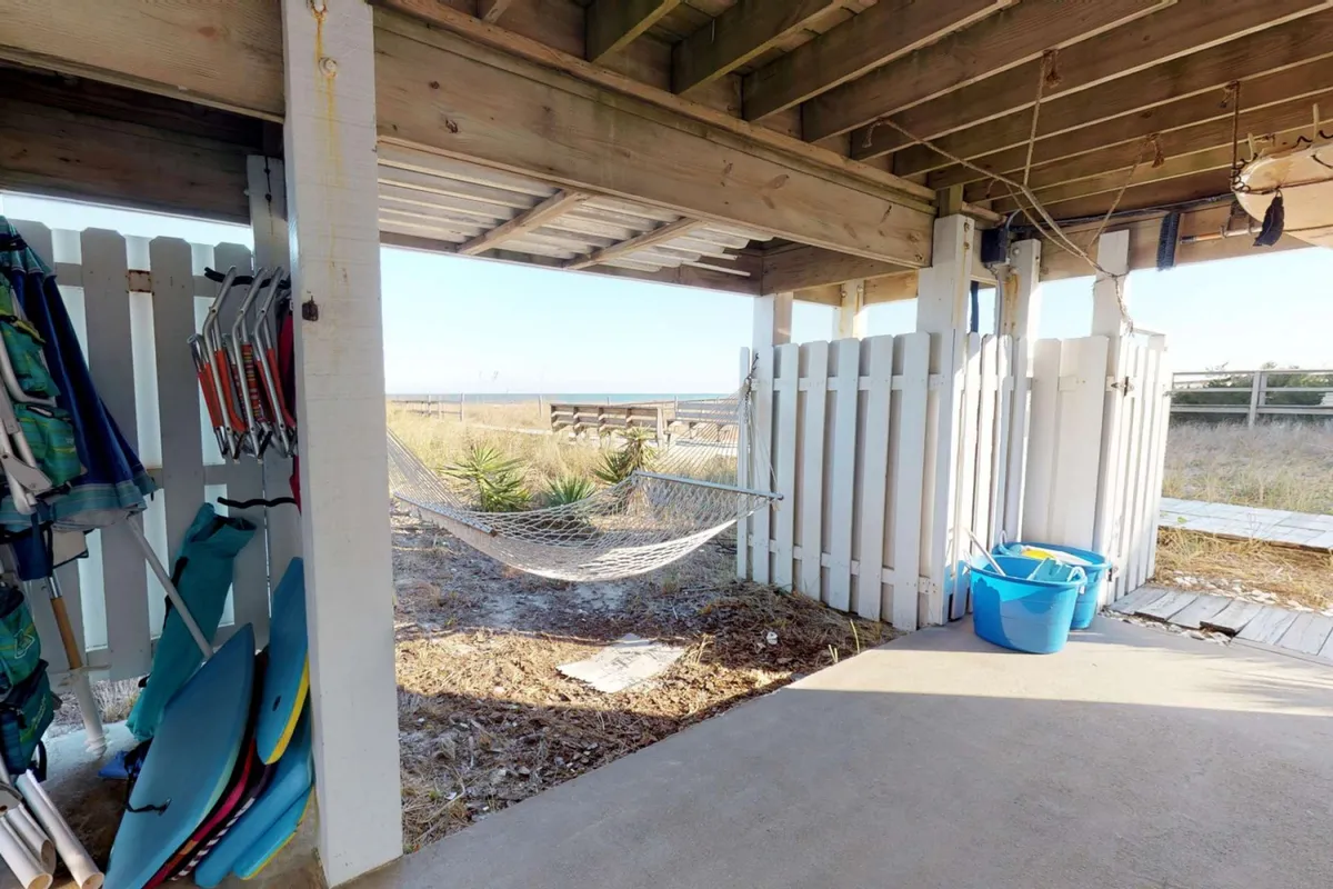 A cozy backyard scene showing a hammock stretched between wooden supports, surfboards propped up nearby, and a blue bucket on the grass. The casual setup encourages outdoor relaxation, reading, and socializing after beach adventures.