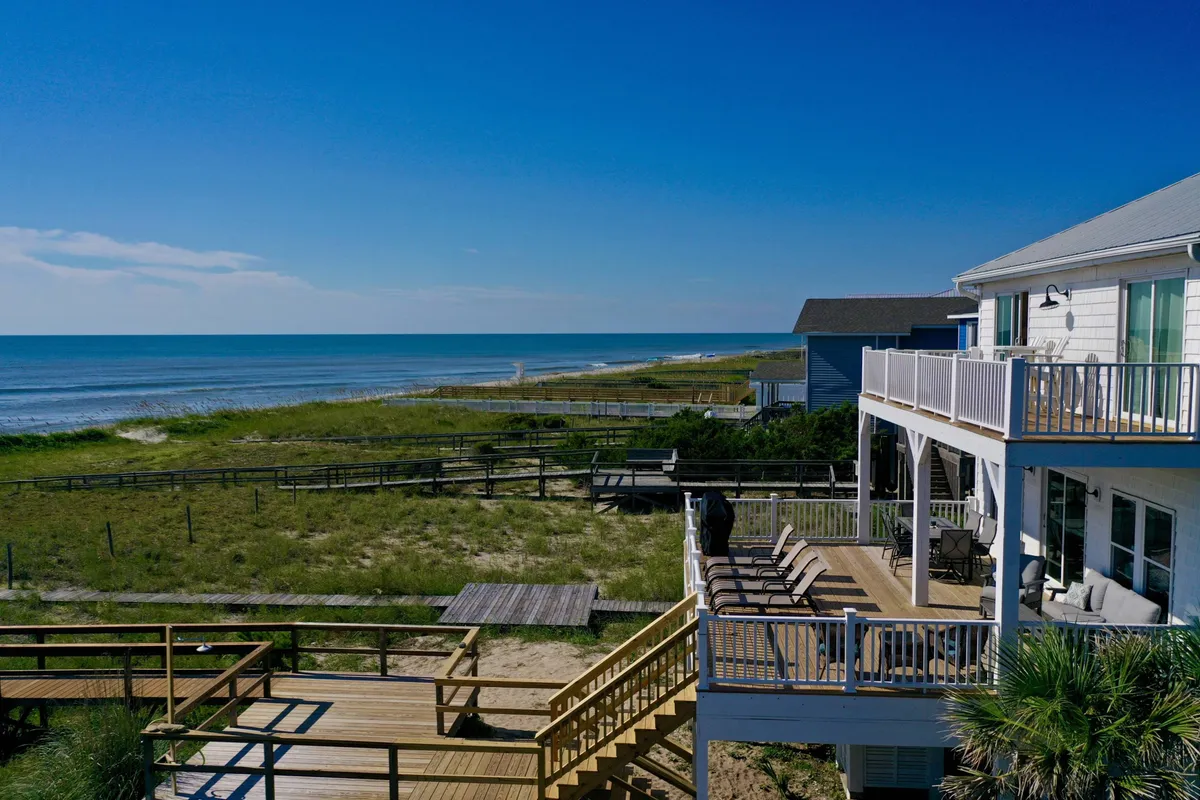 The exterior of a spacious, oceanfront single-family home at Kure Beach, featuring a large deck with inviting seating, lush landscaping, and panoramic views of the beach and Atlantic Ocean. The image highlights the outdoor living space ideal for relaxation, group gatherings, sunrise yoga, sunset dinners, and direct access to the sandy shoreline.