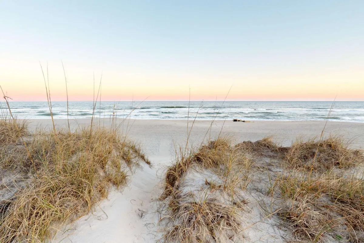 This image showcases sweeping sand dunes with ocean waves rolling in the background and grasses dotting the foreground. The serene landscape offers a sense of openness and natural beauty, inviting viewers to imagine leisurely strolls along the shore.